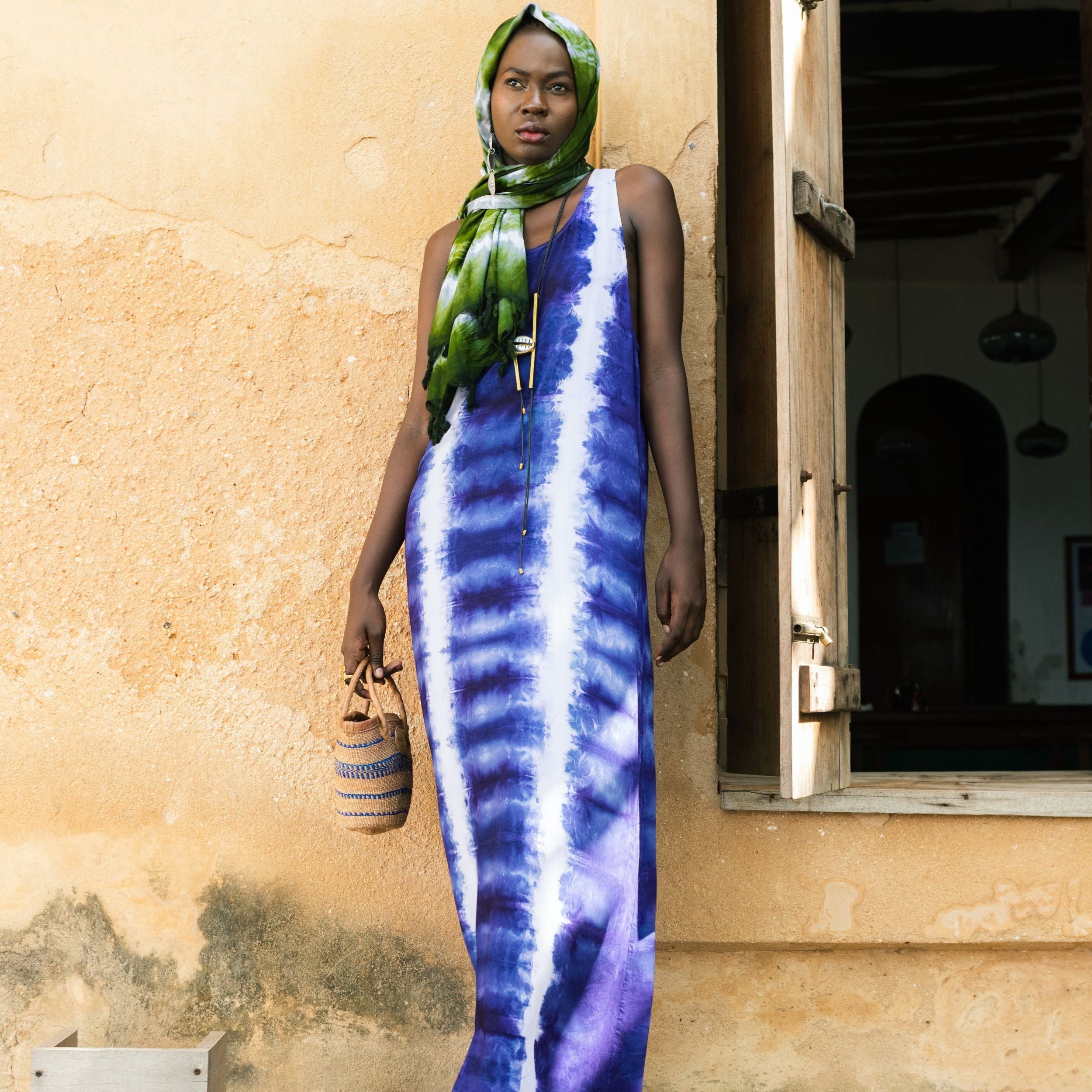 Editorial photo: Model wears Long Dress in Tie Dye Soft Blue while holding a  Mini Shopping Bag in Sisal Blue Stripes and is wearing the Pareo in Tie Dye Intense Green on her head.