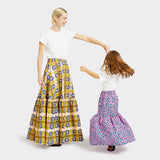 Model, who is a child, wears a Flounced Maxi Skirt Baby in Wax Pretty Little Flowers with colors pink and blue with a plain white tshirt. She is with an adult Model who is wearing a Flounced Maxi Skirt in Wax Magic Mosaic with colors yellow and blue with a plain white shirt. 