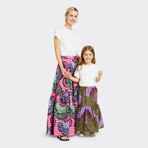 Model, who is a child, wears a Flounced Maxi Skirt Baby in Wax Purple Feathers with colors purple and green with a plain white tshirt. She is with an adult Model who is wearing a Flounced Maxi Skirt in Wax Dreamcatcher in the colors pink, blue, and green with a plain white tshirt. 