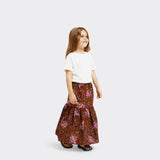 Model who is a child, wears a Flounced Maxi Skirt Baby in Wax Tumeric with colors brown and pink, along with a plain white tshirt.
