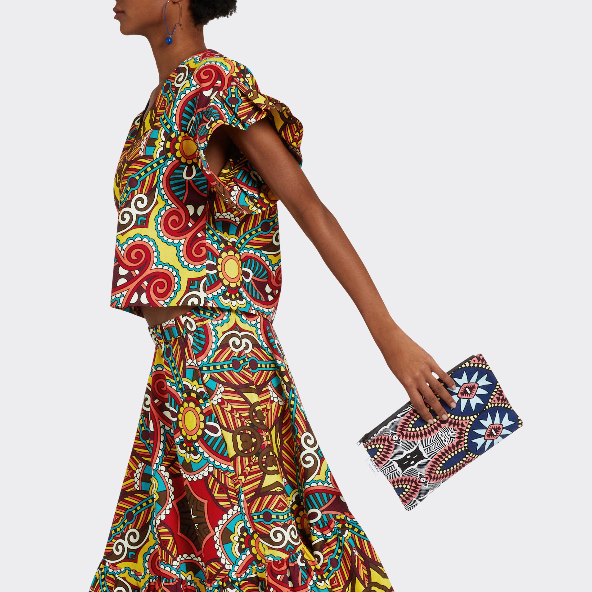 Model is holding the Pouch in Wax Summer Soundtrack which is blue and pink. She wears the Ruffled Top in Wax African Dream with the colors yellow, blue and red and she is wearing the the Flounced Maxi Skirt in Wax African Dream with the colors yellow, blue and red.