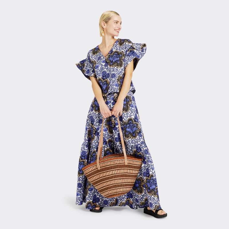 The model is holding the Shopping Bag in Sisal Brown with the color brown. Model wears Ruffled Top in Wax Lilac Peonies with the color lilac and white and the matching Flounced Maxi Skirt in Wax Lilac Peonies in the colors lilac and white for a total look.