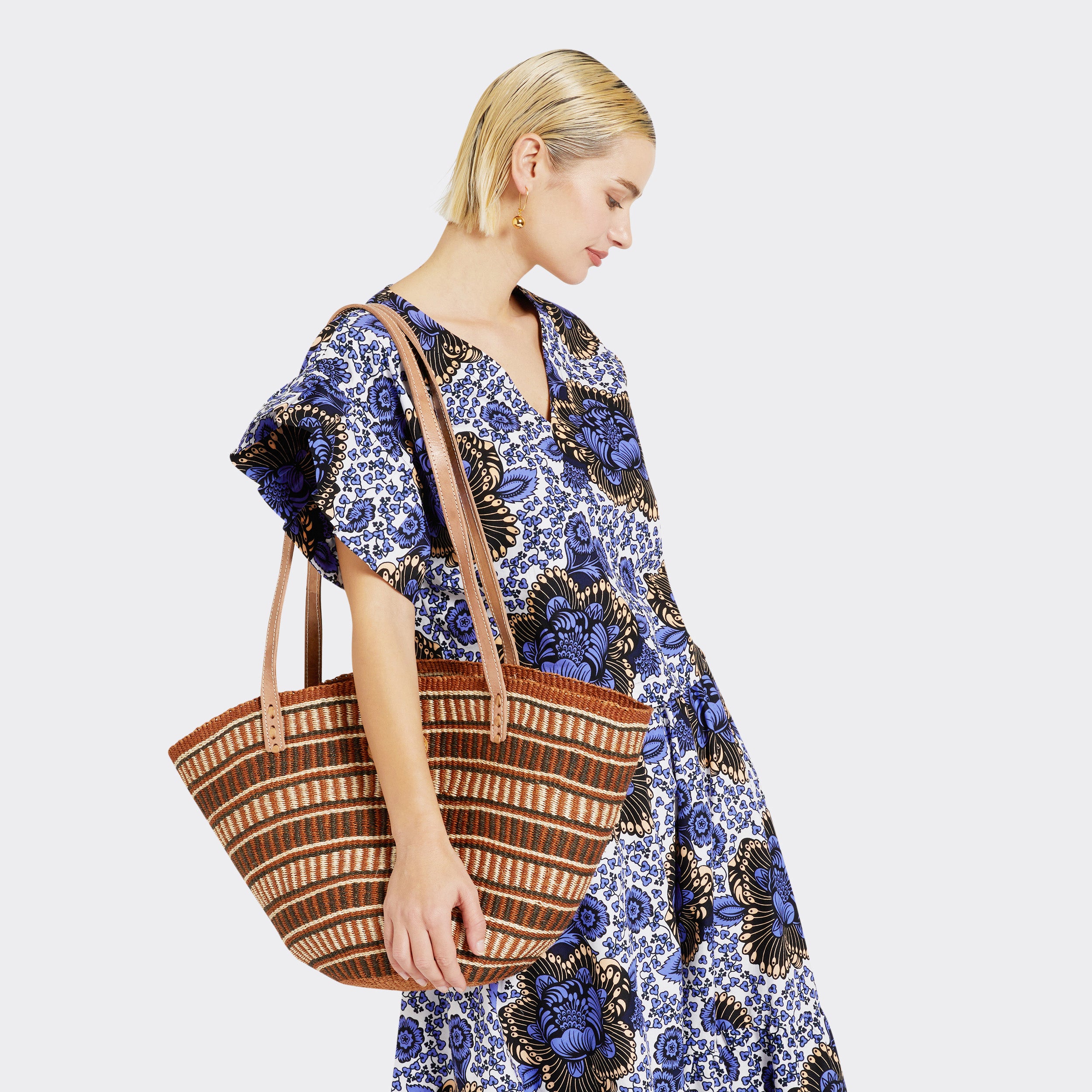The model is holding the Shopping Bag in Sisal Brown with the color brown. Model wears Ruffled Top in Wax Lilac Peonies with the color lilac and white and the matching Flounced Maxi Skirt in Wax Lilac Peonies in the colors lilac and white for a total look.