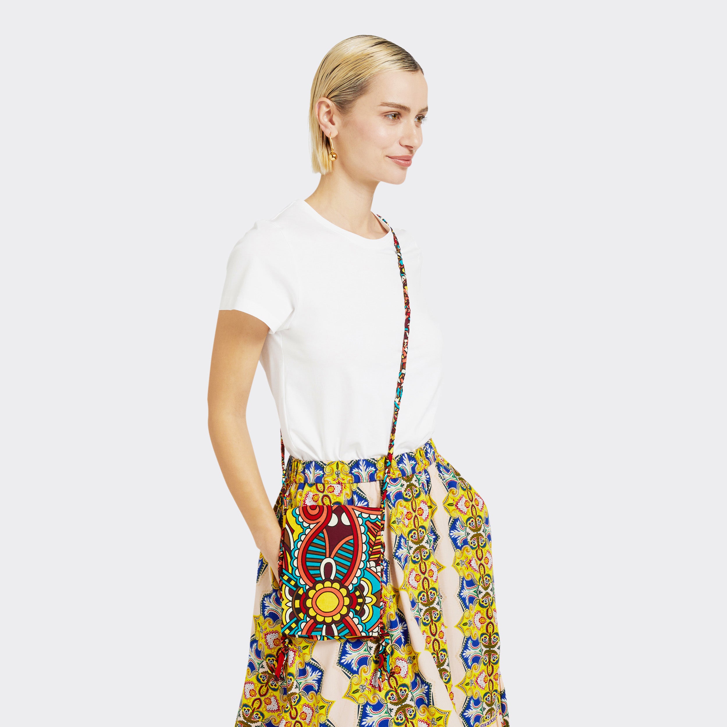 The model is wearing the Crossbody Mini Bag in Wax African Dream with colors red, blue, and yellow. She is wearing the Flounced Maxi Skirt in Wax Magic Mosaic with colors yellow and blue with a plain white tshirt.