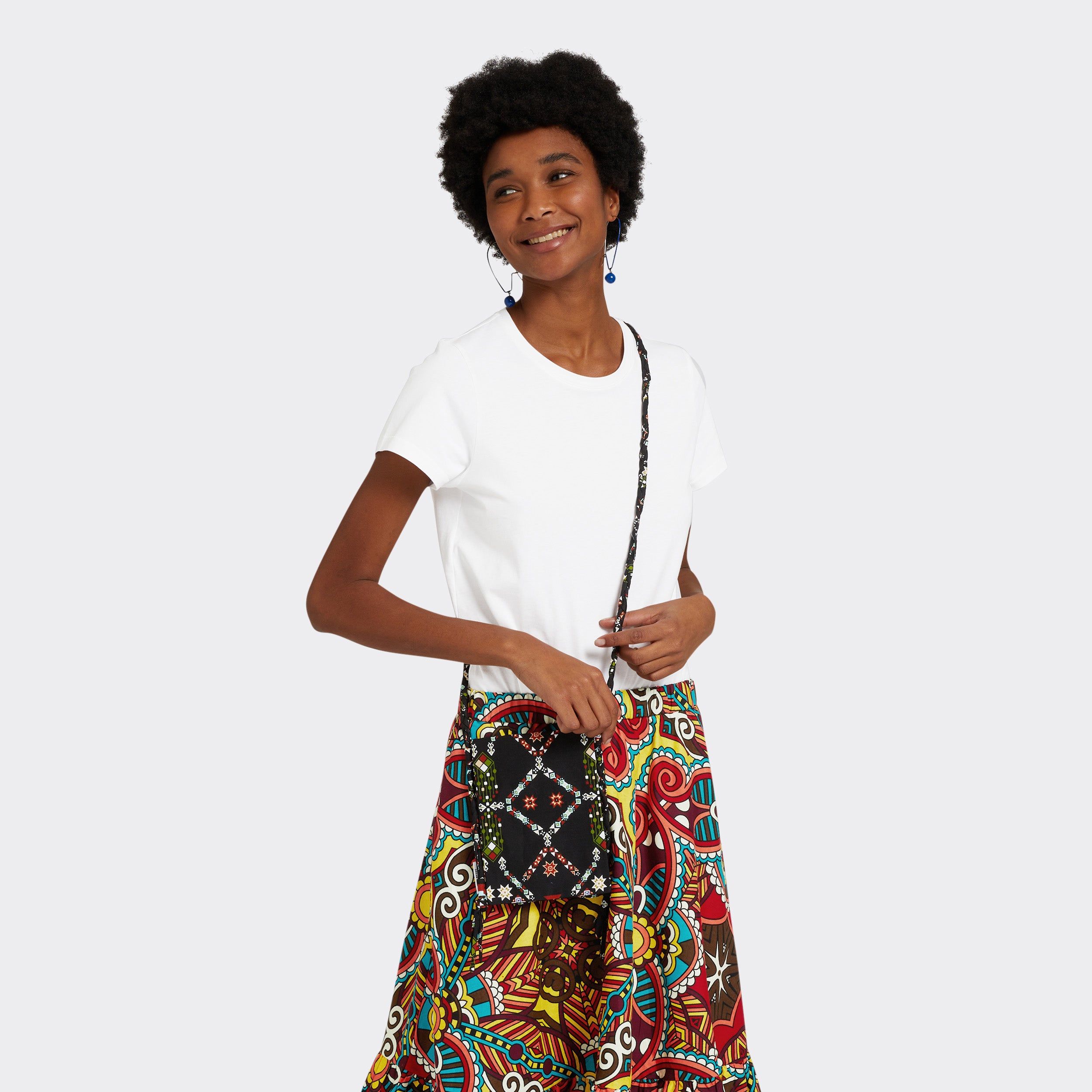 Model is wearing the a Crossbody Mini Bag in Wax Almasi Game with the colors black and red. She is wearing the the Flounced Maxi Skirt in Wax African Dream with the colors yellow, blue and red and a white t shirt.