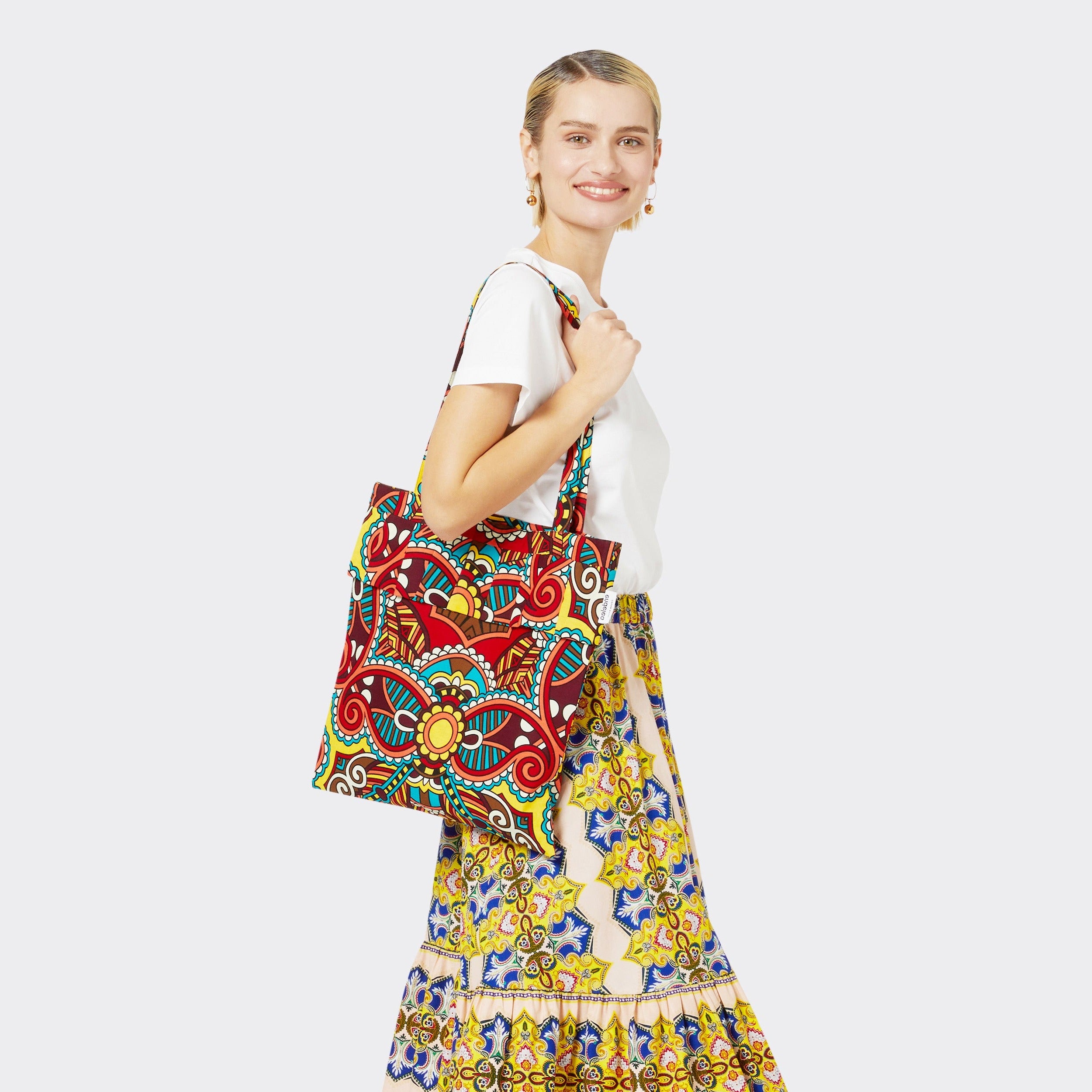 The model is holding the Tote Bag with Rouche in Wax African Dream with colors red, blue, yellow. She is wearing the Flounced Maxi Skirt in Wax Magic Mosaic with colors yellow and blue with a plain white tshirt.