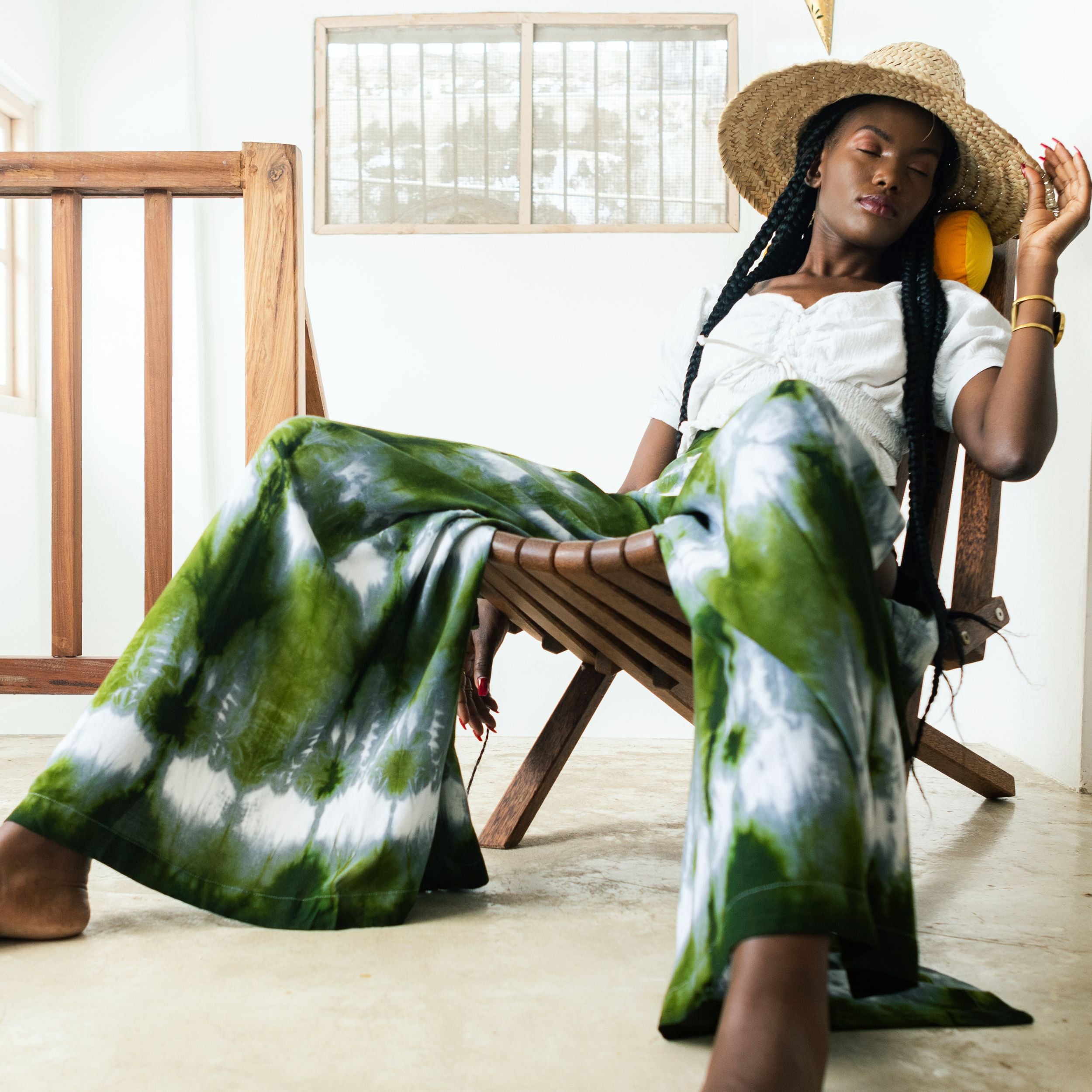 Editorial photo: Model wears Wrap Trousers in Tie Dye Intense Green with a white top and a hat. She is sitting in a wooden chair.